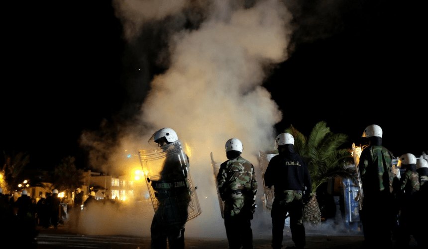 Smoke rises as riot police officers stand guard separating protesting groups of locals and refugees
