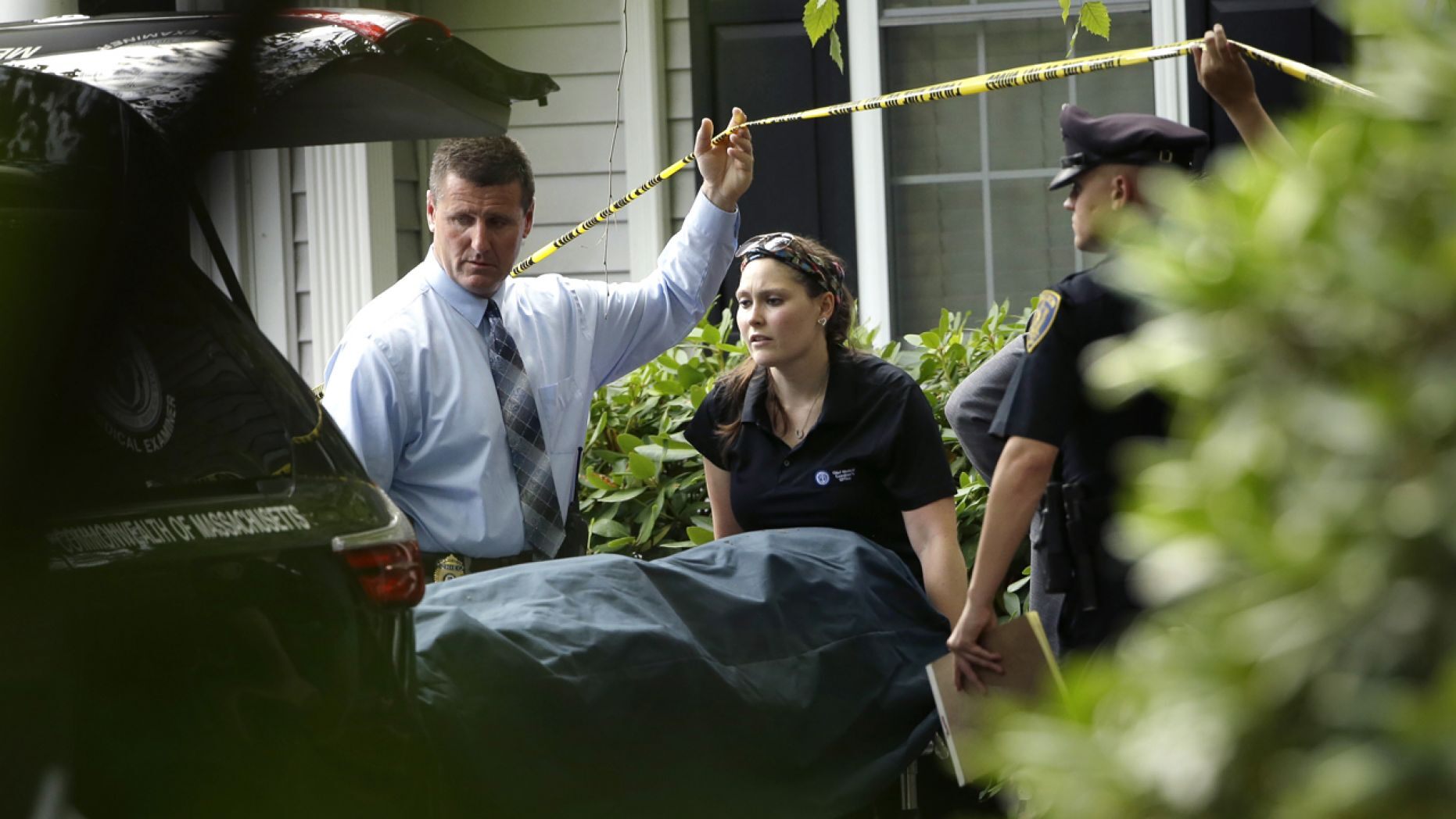 A woman from the Massachusetts Chief Medical Examiners Office, center, uses a gurney to place human remains into a vehicle as law enforcement officers hold caution tape at a home where two adults and three children were found dead with gunshot wounds, Monday, Oct. 7, 2019, in Abington, Mass.