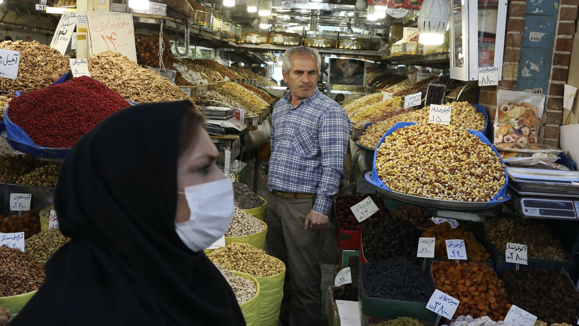 In this photo taken on Tuesday, March 17, 2020, a shopkeeper waits for customer as a woman wearing a face mask to help protect against the new coronavirus, walks at the Tehran's Grand Bazaar, Iran